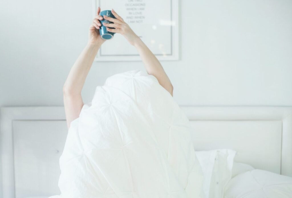 Femme allongée dans un lit aux draps blancs, tenant une tasse en levant les bras au réveil