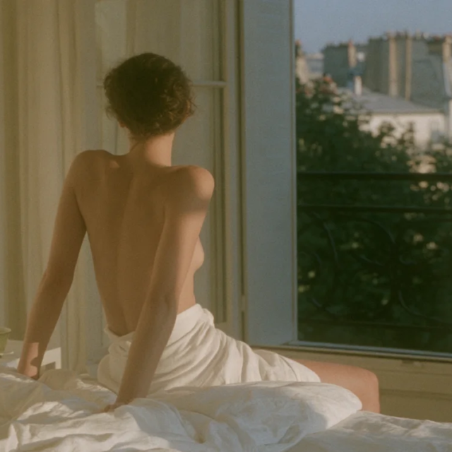Femme assise de dos sur son lit, face à la fenêtre, au réveil